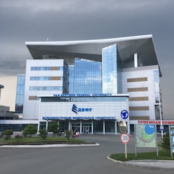 A modern building with a distinctive architectural design featuring large windows and a prominent overhanging roof. The facade is composed of reflective glass panels. It is identified as Far Eastern Federal University. Surrounding the building is a landscaped area with grass and small trees. There are some people near the entrance, and a 30 km/h speed limit sign along the road in front.