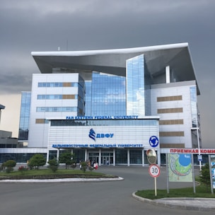 A modern building with a distinctive architectural design featuring large windows and a prominent overhanging roof. The facade is composed of reflective glass panels. It is identified as Far Eastern Federal University. Surrounding the building is a landscaped area with grass and small trees. There are some people near the entrance, and a 30 km/h speed limit sign along the road in front.