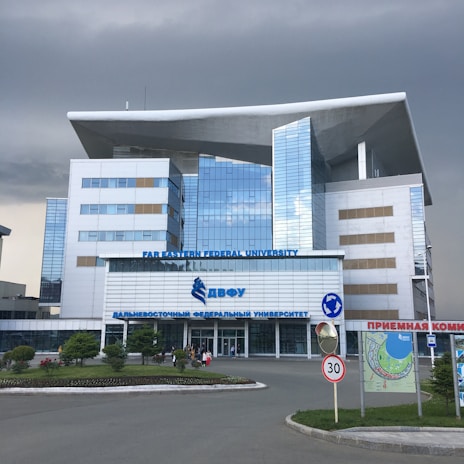 A modern building with a distinctive architectural design featuring large windows and a prominent overhanging roof. The facade is composed of reflective glass panels. It is identified as Far Eastern Federal University. Surrounding the building is a landscaped area with grass and small trees. There are some people near the entrance, and a 30 km/h speed limit sign along the road in front.
