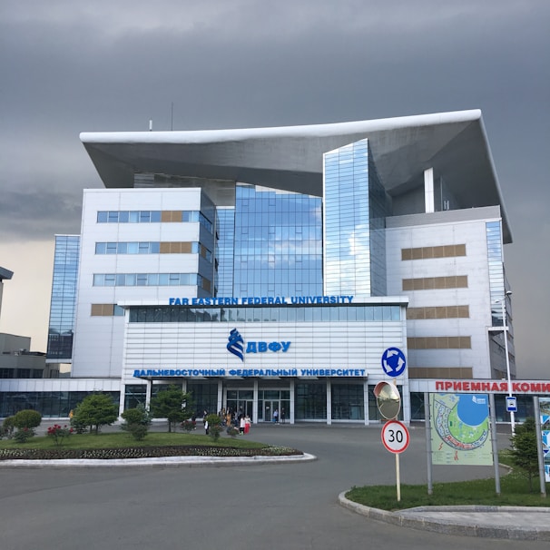 A modern building with a distinctive architectural design featuring large windows and a prominent overhanging roof. The facade is composed of reflective glass panels. It is identified as Far Eastern Federal University. Surrounding the building is a landscaped area with grass and small trees. There are some people near the entrance, and a 30 km/h speed limit sign along the road in front.