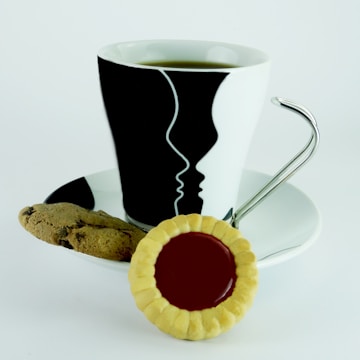 A white cup with artistic black designs depicting faces in profile contains a hot beverage, set on a matching saucer. Next to the cup, there is a chocolate chip cookie and a biscuit with a red jelly center.