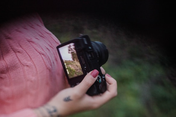 A hand holding a digital camera with the screen displaying a nature scene. The person is wearing a pink knitted sweater, and their nails are painted pink. The background is blurred green foliage, and there is a small tattoo visible on the person's wrist.