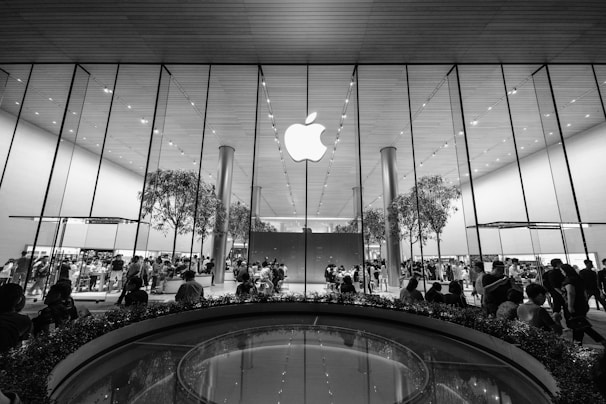 A large, modern retail store with a prominent glass facade featuring a large Apple logo at the center. Inside, numerous people are gathered around tables and displays, with a few trees incorporated within the store design. The ceiling is high with a reflective surface, contributing to the sleek and contemporary aesthetic.