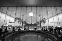 A large, modern retail store with a prominent glass facade featuring a large Apple logo at the center. Inside, numerous people are gathered around tables and displays, with a few trees incorporated within the store design. The ceiling is high with a reflective surface, contributing to the sleek and contemporary aesthetic.