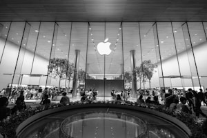 A large, modern retail store with a prominent glass facade featuring a large Apple logo at the center. Inside, numerous people are gathered around tables and displays, with a few trees incorporated within the store design. The ceiling is high with a reflective surface, contributing to the sleek and contemporary aesthetic.