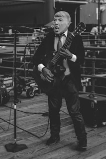 A person wearing a suit and tie is performing with a guitar on a wooden deck, surrounded by microphones and music equipment. The setting appears to be an outdoor venue or street performance.