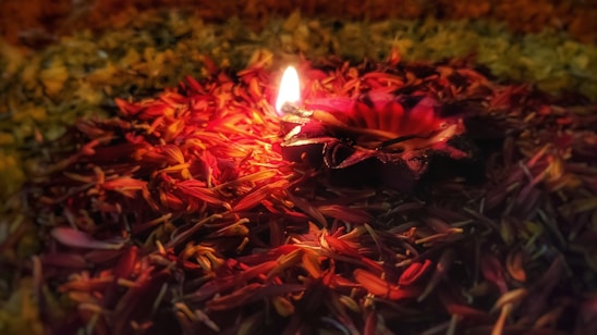 A diya or traditional oil lamp is surrounded by a bed of vibrant red and yellow flower petals, creating a warm and serene atmosphere. The flame casts a soft glow, illuminating the surrounding petals.