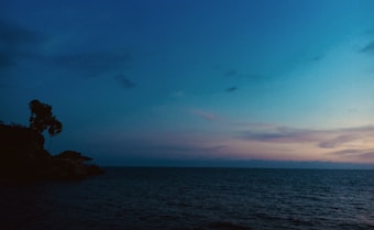 A serene seascape with a soft gradient of colors in the sky, transitioning from deep blue to light purple and orange hues near the horizon. The silhouette of a tree and a small structure stand on a rocky outcrop to the left, with the calm sea extending across the image.