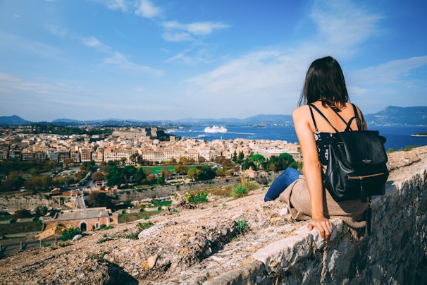 A person with a backpack sits on a stone wall, overlooking a sprawling coastal city. The view includes historic architecture, lush greenery, and a cruise ship in the distance under a bright blue sky.
