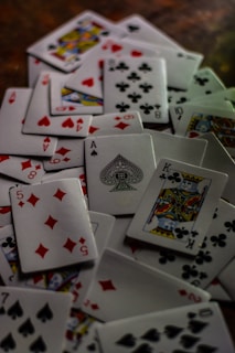 A scattered pile of playing cards featuring various suits and numbers, prominently displaying cards like the King of Clubs, Ace of Spades, and the Five of Diamonds. The cards are randomly distributed creating a chaotic yet intriguing arrangement.