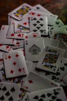 A scattered pile of playing cards featuring various suits and numbers, prominently displaying cards like the King of Clubs, Ace of Spades, and the Five of Diamonds. The cards are randomly distributed creating a chaotic yet intriguing arrangement.