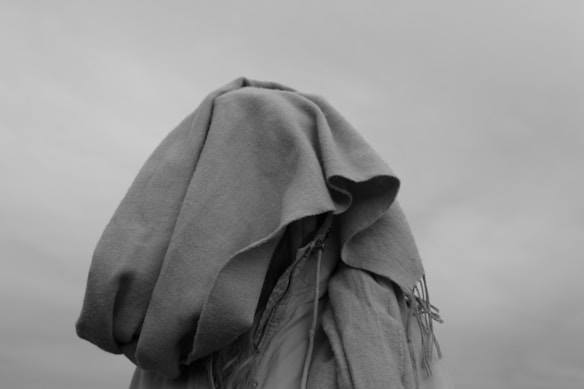 A person with their head covered by a large, draped cloth, creating a mysterious and somewhat anonymous appearance. The image is in black and white, with minimal background details visible.