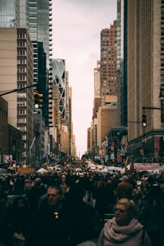 A bustling city street scene with tall skyscrapers lining the sides, creating a canyon-like effect. A large crowd of people is seen walking and engaging with various street vendors and food stalls. The atmosphere feels lively and vibrant, typical of an urban environment filled with diverse activities.