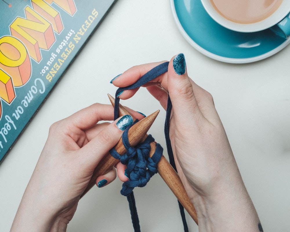 Persona che lega il merletto blu su due bastoncini marroni accanto all'alzavola e al set di tazze da tè in ceramica bianca