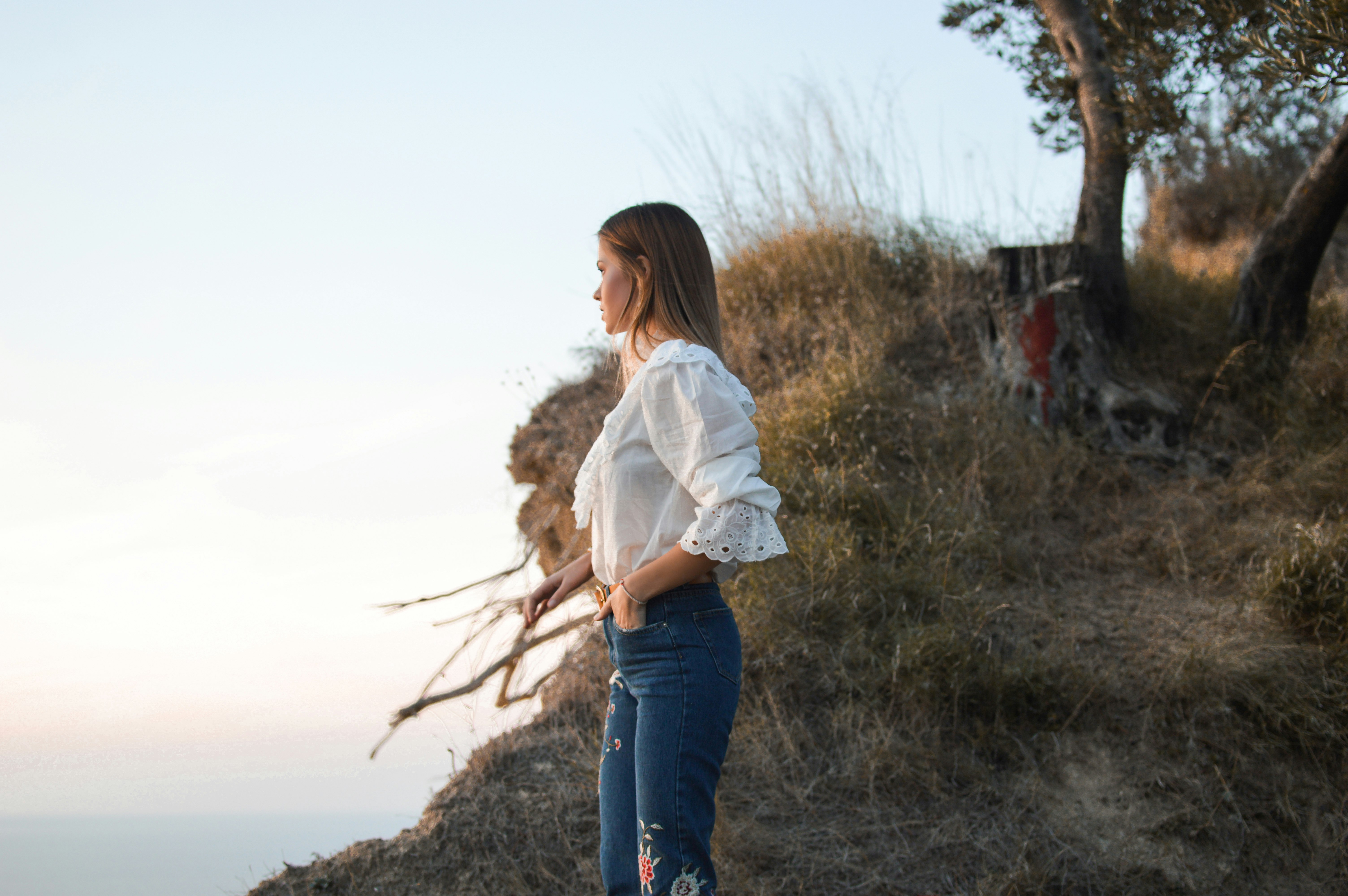 woman standing in mountain wearing white blouse watching body of water during daytime