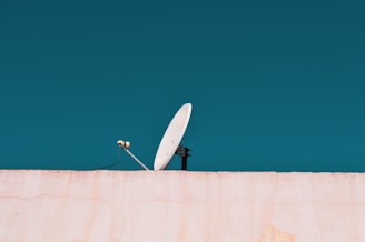 A white satellite dish is mounted on a light-colored wall against a clear cyan sky. The dish is tilted slightly upwards and supported by a metal arm.