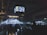 A dimly lit indoor concert venue with a large crowd facing a stage. Bright lights and spotlights illuminate the area, with screens hanging from the ceiling displaying video feeds. The atmosphere is lively, with people standing and some sitting on the floor, enjoying the music.