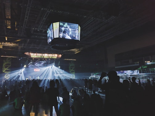 A dimly lit indoor concert venue with a large crowd facing a stage. Bright lights and spotlights illuminate the area, with screens hanging from the ceiling displaying video feeds. The atmosphere is lively, with people standing and some sitting on the floor, enjoying the music.