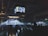 A dimly lit indoor concert venue with a large crowd facing a stage. Bright lights and spotlights illuminate the area, with screens hanging from the ceiling displaying video feeds. The atmosphere is lively, with people standing and some sitting on the floor, enjoying the music.