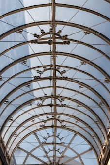 A glass roof with an intricate metal framework featuring circular and geometric designs. Sunlight filters through, creating a pattern of shadows and light on the surface.