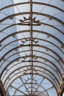 A glass roof with an intricate metal framework featuring circular and geometric designs. Sunlight filters through, creating a pattern of shadows and light on the surface.