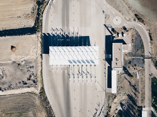 An aerial view of a toll plaza situated on a wide, empty road with several lanes. The toll booths are lined up in a neat row under a large white canopy, casting long shadows on the ground. The surrounding area includes dry, barren land with some vegetation and a few structures, along with roadways leading in different directions.