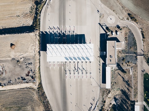 An aerial view of a toll plaza situated on a wide, empty road with several lanes. The toll booths are lined up in a neat row under a large white canopy, casting long shadows on the ground. The surrounding area includes dry, barren land with some vegetation and a few structures, along with roadways leading in different directions.