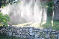 Water sprinklers creating a misty effect over a garden, with sunlight streaming through the water droplets. A stone wall is in the foreground, and lush green vegetation surrounds the scene.
