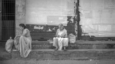 Two women are sitting on a stone ledge in front of a wall with torn posters. One woman looks contemplative while the other is focused on something in her hands. Both are dressed in traditional attire, and there are bags placed next to them.