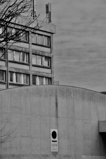 A monochrome image featuring a large concrete building with multiple windows, an antenna on the rooftop, and a branch from a leafless tree in the foreground. A sign with the letters 'UCO' is prominently displayed on the front of the building.