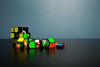 A broken Rubik's Cube with colorful pieces scattered on a dark surface against a plain, teal background. The cube is primarily black with visible green and yellow tiles, and the disassembled pieces include red and blue colors.