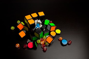 A disassembled Rubik's cube lies on a dark surface, surrounded by colorful plastic pieces and small parts. The cube's core is exposed, revealing a blue mechanism at the center, while various stickers in orange, green, and other colors are visible.