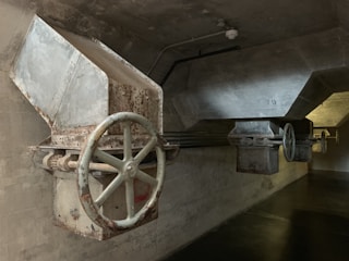 A dimly lit industrial setting featuring large metal hoppers or chutes with visible rust and wear. The structures have big, circular valves or wheels attached, mounted on a concrete wall and ceiling. The lighting is subdued, casting shadows on the walls and floor.