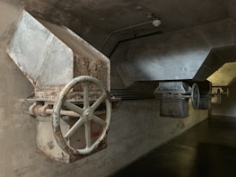 A dimly lit industrial setting featuring large metal hoppers or chutes with visible rust and wear. The structures have big, circular valves or wheels attached, mounted on a concrete wall and ceiling. The lighting is subdued, casting shadows on the walls and floor.