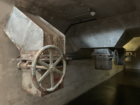 A dimly lit industrial setting featuring large metal hoppers or chutes with visible rust and wear. The structures have big, circular valves or wheels attached, mounted on a concrete wall and ceiling. The lighting is subdued, casting shadows on the walls and floor.