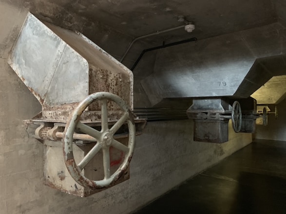 A dimly lit industrial setting featuring large metal hoppers or chutes with visible rust and wear. The structures have big, circular valves or wheels attached, mounted on a concrete wall and ceiling. The lighting is subdued, casting shadows on the walls and floor.