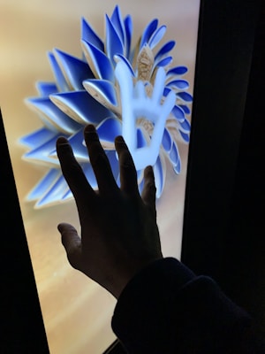 A hand is reaching out toward a bright screen displaying a digital artwork. The artwork consists of blue and white spiky shapes resembling feathers or petals, with a glowing hand symbol in the center making a 'rock on' gesture.
