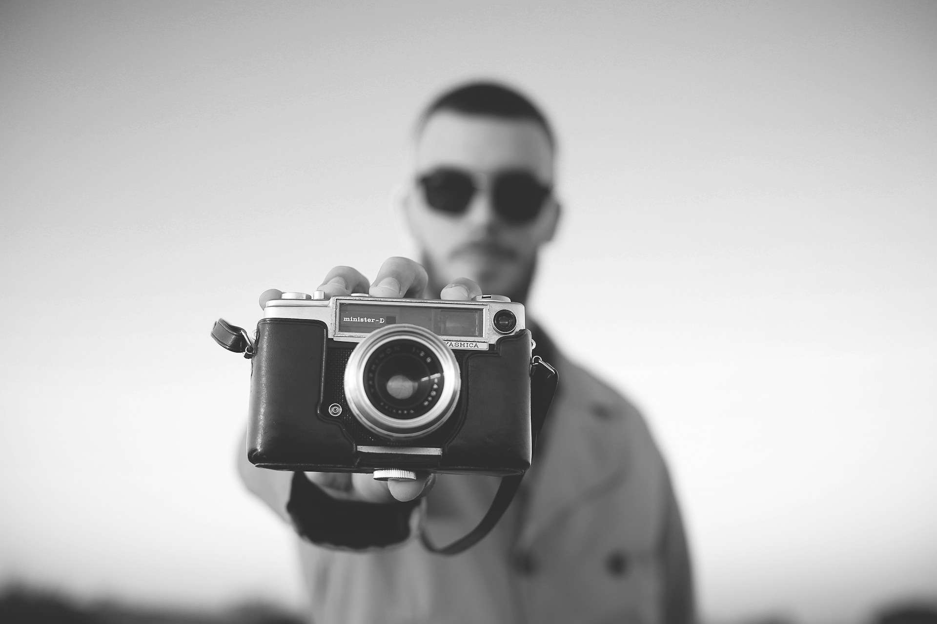 A person holding a vintage camera in the foreground, with the focus on the camera. The individual is blurred in the background, wearing sunglasses and a coat. The image is in black and white, adding a classic and timeless feel.