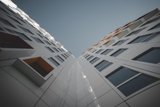 The image depicts a modern architectural design featuring two tall, adjacent buildings showcasing a facade with geometric patterns and large windows. The perspective is upward, creating a dramatic vanishing point that emphasizes the height and symmetry of the structures. Subtle accents in the design include colorful elements near the top of the buildings, possibly serving as decorative panels or sun shades.
