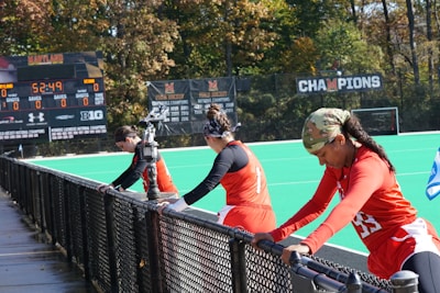 A group of athletes wearing orange uniforms are on the sideline of a sports field, resting against a fence. Behind them, a large digital scoreboard displays team names and scores. The field is bright green, and several banners with sponsor logos and championship wins are visible. The athletes appear to be taking a break, possibly before or during a field hockey game.