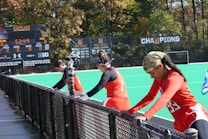 A group of athletes wearing orange uniforms are on the sideline of a sports field, resting against a fence. Behind them, a large digital scoreboard displays team names and scores. The field is bright green, and several banners with sponsor logos and championship wins are visible. The athletes appear to be taking a break, possibly before or during a field hockey game.