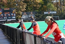 A group of athletes wearing orange uniforms are on the sideline of a sports field, resting against a fence. Behind them, a large digital scoreboard displays team names and scores. The field is bright green, and several banners with sponsor logos and championship wins are visible. The athletes appear to be taking a break, possibly before or during a field hockey game.