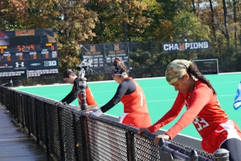 A group of athletes wearing orange uniforms are on the sideline of a sports field, resting against a fence. Behind them, a large digital scoreboard displays team names and scores. The field is bright green, and several banners with sponsor logos and championship wins are visible. The athletes appear to be taking a break, possibly before or during a field hockey game.