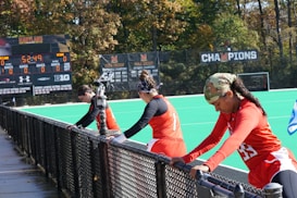 A group of athletes wearing orange uniforms are on the sideline of a sports field, resting against a fence. Behind them, a large digital scoreboard displays team names and scores. The field is bright green, and several banners with sponsor logos and championship wins are visible. The athletes appear to be taking a break, possibly before or during a field hockey game.