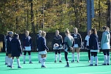 A group of people, primarily young women, wearing athletic uniforms and jackets are standing in a circle on a green sports field. One person is in the center of the circle, appearing to balance a ball on their knee. The background features trees with autumn foliage.