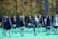 A group of people, primarily young women, wearing athletic uniforms and jackets are standing in a circle on a green sports field. One person is in the center of the circle, appearing to balance a ball on their knee. The background features trees with autumn foliage.