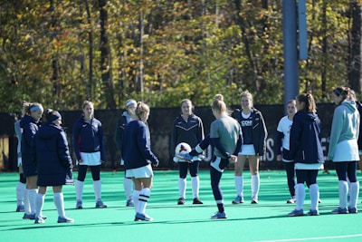 A group of people, primarily young women, wearing athletic uniforms and jackets are standing in a circle on a green sports field. One person is in the center of the circle, appearing to balance a ball on their knee. The background features trees with autumn foliage.