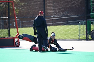 Two individuals in field hockey gear are on an outdoor sports field. One person is in a goalie uniform, sitting on the ground with a hockey stick in hand, while the other stands nearby, wearing a cap and observing. A goalpost with a net is positioned on the left side.