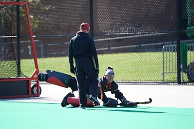 Two individuals in field hockey gear are on an outdoor sports field. One person is in a goalie uniform, sitting on the ground with a hockey stick in hand, while the other stands nearby, wearing a cap and observing. A goalpost with a net is positioned on the left side.