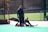 Two individuals in field hockey gear are on an outdoor sports field. One person is in a goalie uniform, sitting on the ground with a hockey stick in hand, while the other stands nearby, wearing a cap and observing. A goalpost with a net is positioned on the left side.